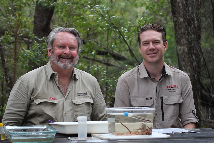 Two men smiling at the camera with a tank containing what looks like a small eel in front of them